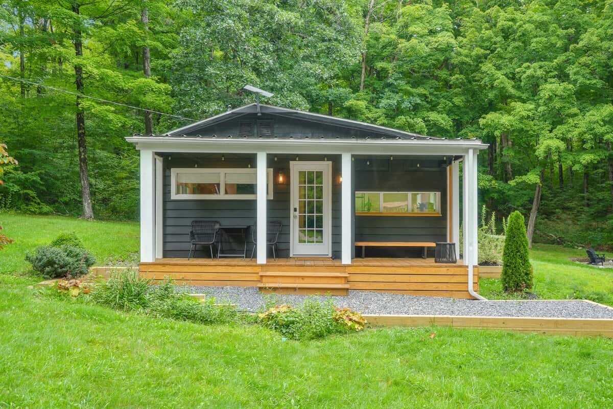 The cabin straight on from the lawn: black board-and-batten siding, white porch posts, a cedar deck, and the forest rising behind the roofline
