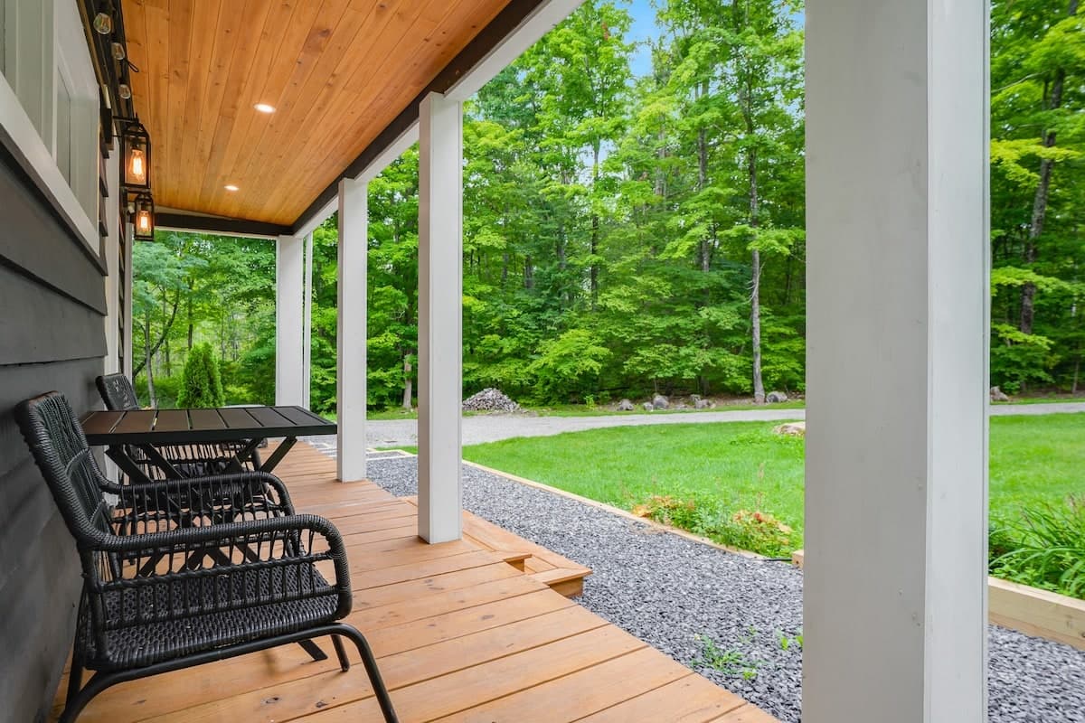 The covered porch: a cedar-plank ceiling, a black table and two chairs, the gravel apron and lawn beyond