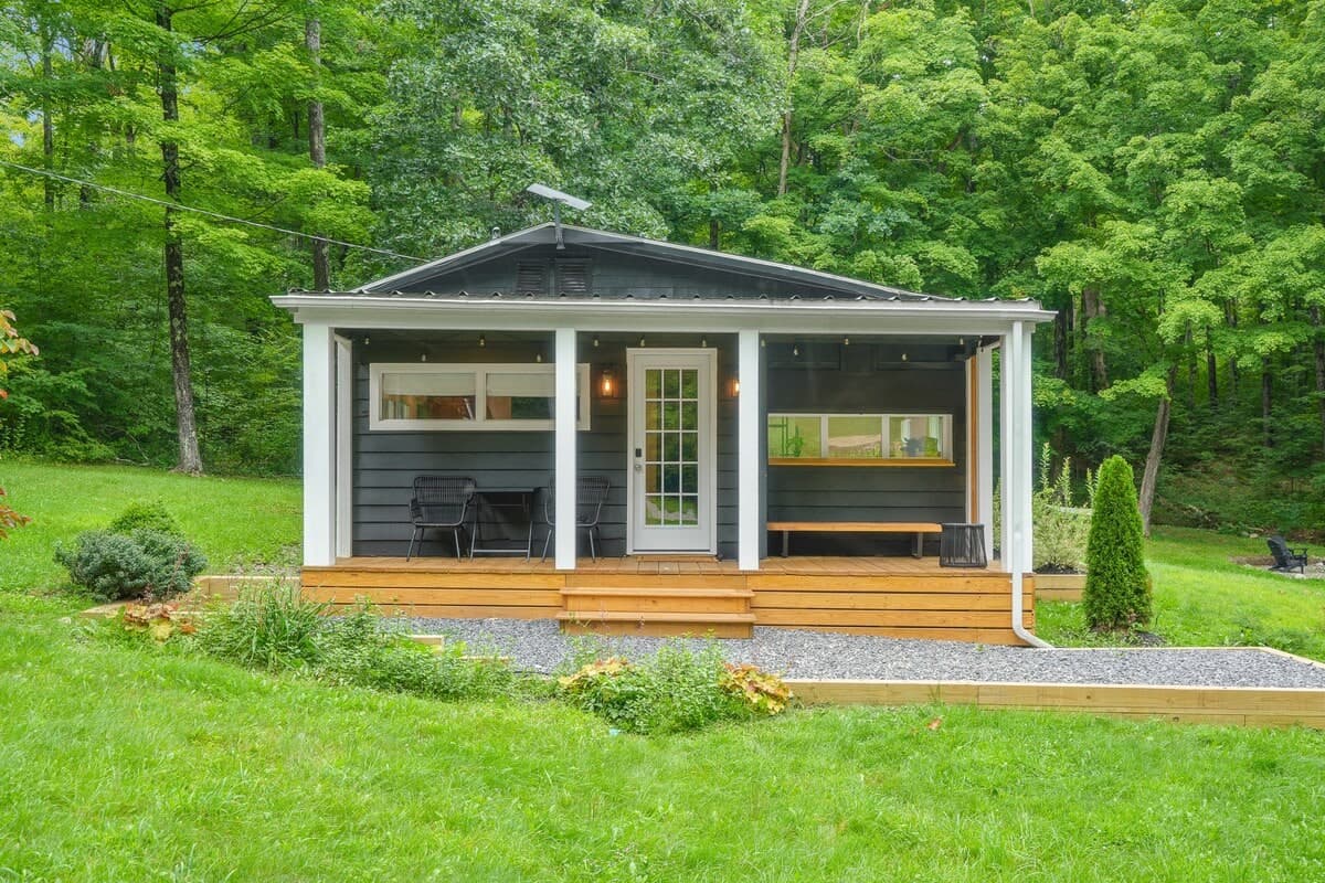 The cabin straight on from the lawn: black board-and-batten siding, white porch posts, a cedar deck, and the forest rising behind the roofline